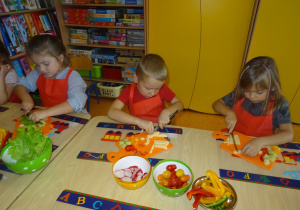 Troje dzieci siedzi przy stole, w ręku trzymają nóż którym kroją z zaangażowaniem sałatę i żółty ser. Na środku stołu stoją miseczki z warzywami.