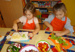 Dwoje dzieci siedzi przy stole, w ręku trzymają nóż którym kroją z zaangażowaniem sałatę i ogórka. Na środku stołu stoją miseczki z warzywami.
