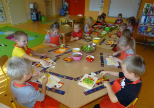 Grupa dzieci siedzi przy dużym stole, trzymają w ręku noże którymi kroją owoce, warzywa oraz ser, przed nimi są rozłożone deseczki a na środku stołu stoją miski z warzywami i owocami.