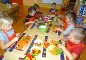 Siedmioro dzieci siedzi przy stole, troje z nich trzyma w ręku nóż i kroi warzywa. Czworo dzieci w ręku trzyma patyczek na który nakładają pokrojone warzywa i owoce.