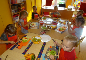 Ośmioro dzieci siedzi przy stole, troje z nich trzyma w ręku noz, którym kroją na deseczkach warzywa. Reszta dzieci nakłada na patyczki przygotowane warzywa i owoce.