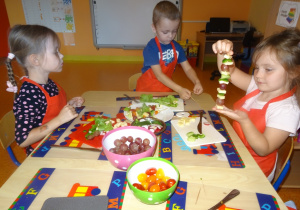 Troje dzieci siedzi przy stole. Jedna z dziewczynek prezentuje przygotowanego szaszłyka owocowo- warzywnego. Chłopiec podnosi ze stolika patyczki do szaszłyków. Druga z dziewczynek obserwuje co robi chłopiec.