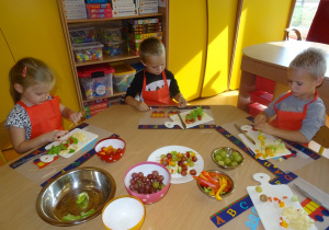 Troje dzieci siedzi przy stole i nakłada warzywa i owoce na patyczki.