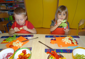 Dwoje dzieci siedzi przy stole i nakłada pomidora oraz sałatę na patyczki.