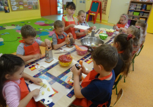 Grupa dzieci siedzi przy dużym stole, dzieci trzymają w ręku tarkę i marchewkę, którą ścierają. Jedna z dziewczynek ma przed sobą deseczkę na której kroi nożem gruszkę.
