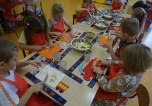 Grupa dzieci siedzi przy dużym stole w ręku trzymają noże którymi kroją gruszki i jabłka na deseczkach.
