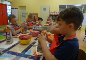 Grupa dzieci siedzi przy dużym stole, dzieci trzymają w ręku tarkę i marchewkę, którą ścierają.