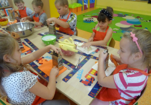 Sześcioro dzieci siedzi przy stole. Pięcioro dzieci trzyma w ręku tarkę na której ściera marchewkę, Jedna dziewczynka ma przed sobą deseczkę na której kroi nożem jabłko.