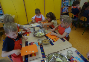 Pięcioro dzieci siedzi przy stole i kroi nożem na desce owoce. Jeden z chłopców unosi deskę z pokrojonym owocem.