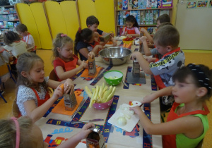 Grupa dzieci siedzi przy stole, w ręku trzymają tarki na których ścierają marchewkę. Jedna z dziewczynek kroi nożem owoc na desce.