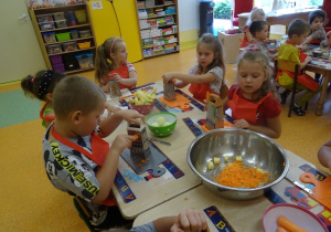 Pięcioro dzieci siedzi przy stole i ściera na tarce marchewkę. Na środku stołu stoi miska z startą marchewką.