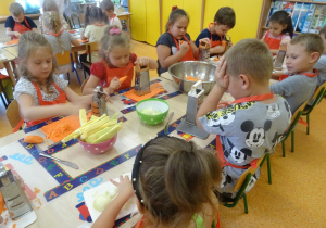Grupa dzieci siedzi przy stole i ściera marchewkę na tarce. Jedna z dziewczynek kroi owoc.