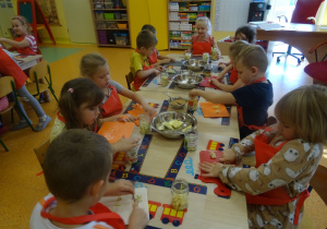 Grupa dzieci siedzi przy stole przed nimi leżą deseczki i słoiki. Część z dzieci kroi nożem owoce a reszta z nich wypełnia pokrojonymi owocami słoiki.