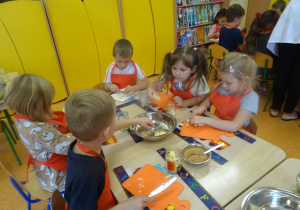 Pięcioro dzieci siedzi przy stole, w ręku trzymają noże którymi kroją owoce na deskach.