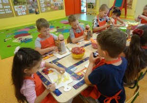 Grupa dzieci siedzi przy dużym stole, dzieci trzymają w ręku tarkę i marchewkę, którą ścierają. Jedna z dziewczynek ma przed sobą deseczkę na której kroi nożem gruszkę.