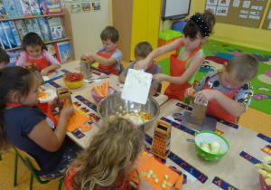 Grupa dzieci siedzi przy stole i ściera marchewkę na tarce. Jedna z dziewczynek stoi przy stole i zrzuca z deski do miski na środku stołu pokrojone owoce.