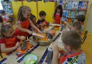 Grupa dzieci siedzi przy stole i ściera marchewkę, jedna z dziewczynek stoi i zrzuca z deski pościeraną marchewkę do miski na środku stołu.