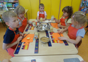 Pięcioro dzieci siedzi przy stole, przed każdym z dzieci stoją wypełnione owocami słoiczki.