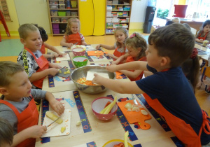 Sześcioro dzieci siedzi przy stole, w ręku trzymają noże którymi kroją owoce. Chłopiec stoi przy stole i zrzuca z deski pokrojone warzywa.