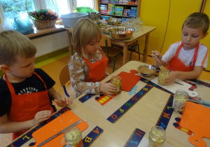 Troje dzieci siedzi przy stole przed sobą mają słoiki wypełnione owocami. Jeden z chłopców sypie łyżką cukier do słoika.