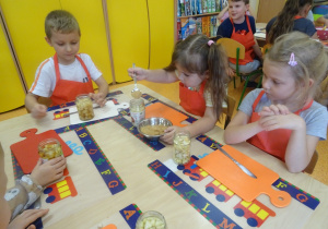 Troje dzieci siedzi przy stole przed sobą mają słoiki wypełnione owocami. Jedna z dziewczynek sypie łyżką cukier do słoika.