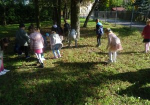 Dzieci zbierają kasztany z trawy, w tle widać drzewa.