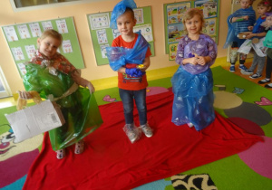 Troje dzieci w ekologicznych strojach stoi na czerwonym dywanie.