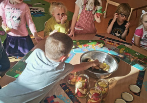dzieci siedzą przy stoliku i jedzą jabłka, które im zostały. Widać przygotowane słoiki z jabłkami