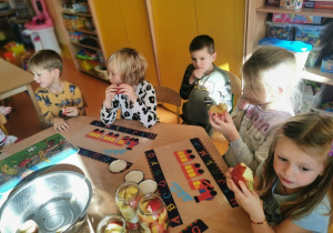 dzieci siedzą przy stoliku jedzą jabłka. Widać przygotowane słoiki z jabłkami