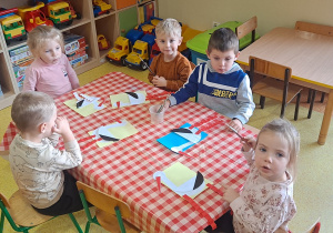 dzieci przy stolikach przyklejają przygotowane przez nauczyciela elementy bociana (tułów, szyja, głowa, dziób, długie czerwone nogi)