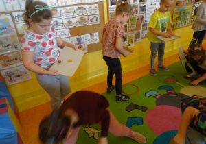 dzieci na dywanie wykonują ćwiczenia na dywanie - ich zadaniem jest tak ruszanie tekturą aby przez zrobioną w niej dziurę przeleciał kasztan i wylądował i obręczy leżącej na dywanie