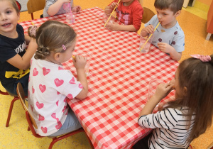 dzieci przy stolikach dmuchają w kubki z woda słomkami