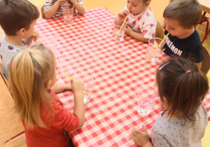 dzieci przy stolikach dmuchają w kubki z woda słomkami
