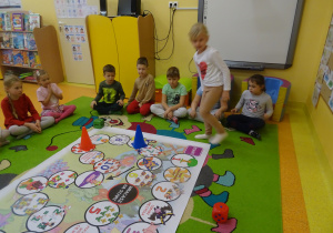 dzieci na dywanie grają w planszówkę, rzucają kostką poruszają się odpowiednią ilość oczek, wykonują zadanie o wylosowanego pola