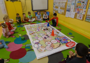 dzieci na dywanie grają w planszówkę, rzucają kostką poruszają się odpowiednią ilość oczek, wykonują zadanie o wylosowanego pola