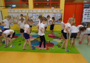dzieci na dywanie wykonują ćwiczenia wskazane przez nauczyciela chodzą z woreczkami na stopach