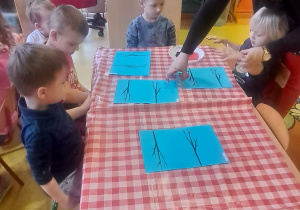 dzieci odciskają rączki w talerzyku z farbą i odbijają ją na kartce przy gałązkach tworząc gil siedzącego na gałązce
