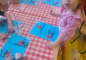 dzieci odciskają rączki w talerzyku z farbą i odbijają ją na kartce przy gałązkach tworząc gil siedzącego na gałązce