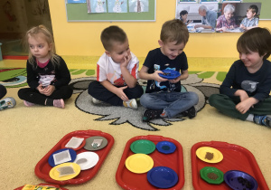 dzieci siedzą na dywanie podają sobie talerzyki i je wąchają
