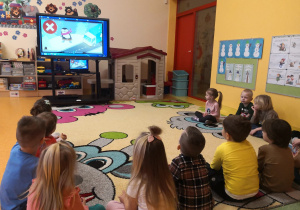 dzieci siedzą po turecku i w uwaga oglądają film edukacyjny o zabawie blisko jezdni