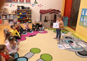 dziewczynka trzyma w rękach ilustracje i pokazuje ją reszcie dzieci siedzącym na dywanie