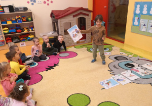 chłopiec trzyma w rękach ilustracje i pokazuje ją reszcie dzieci siedzącym na dywanie