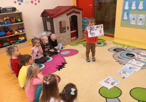 chłopiec pokazuje ilustracje dzieciom siedzącym na dywanie
