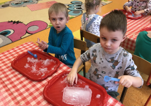 dzieci przy stolikach z bliska obserwują i dotykają bryły śniegu