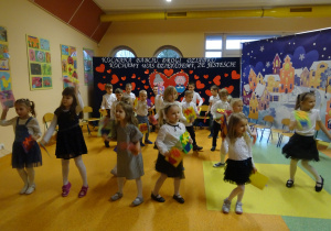 dzieci trzymają kolorowe laurki w rękach i tańczą z nimi dla swoich dziadków
