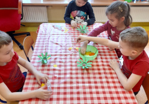 dzieci przy stolikach na końcach gałązek przyklejają kolorowe kwiaty z papieru