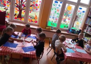 dzieci przy stolikach na niebieskich kartkach przyklejają zielone gałązki