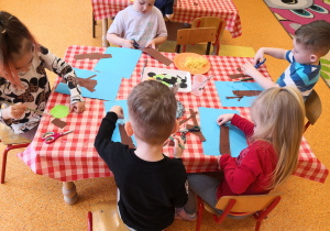 dzieci przy stolikach naklejają gotowe elementy na niebieską kartkę tworząc drzewo