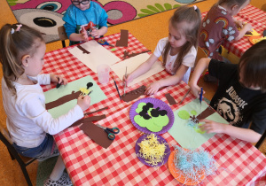 dzieci przy stolikach naklejają gotowe elementy na niebieską kartkę tworząc drzewo