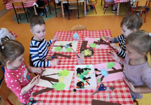 dzieci przy stolikach naklejają gotowe elementy na niebieską kartkę tworząc drzewo oraz gniazdo na nim się znajdujące